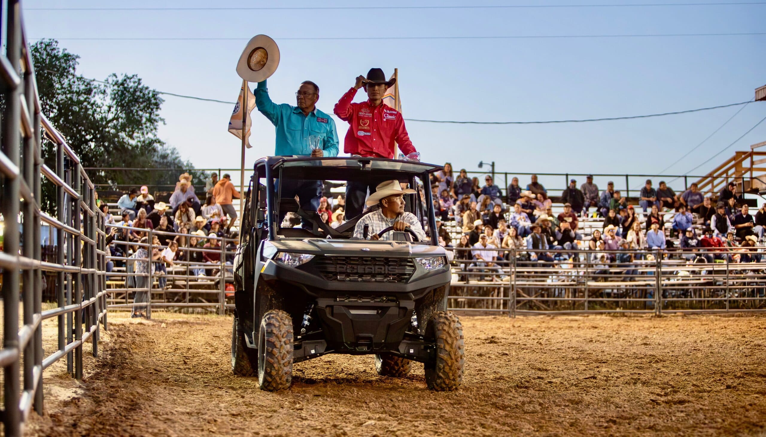 Officials ride in an ATV through a rodeo arena during a parade lap, waving to spectators in the stands