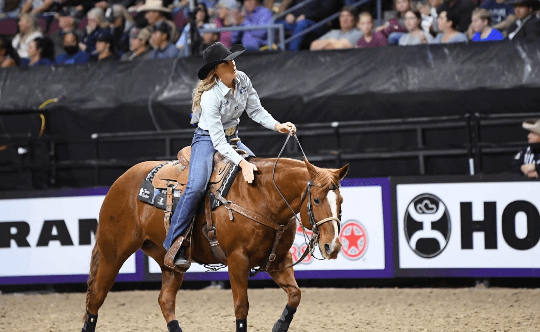 Cowgirl in a black hat and light blue shirt pats her sorrel horse's neck while riding across an indoor arena