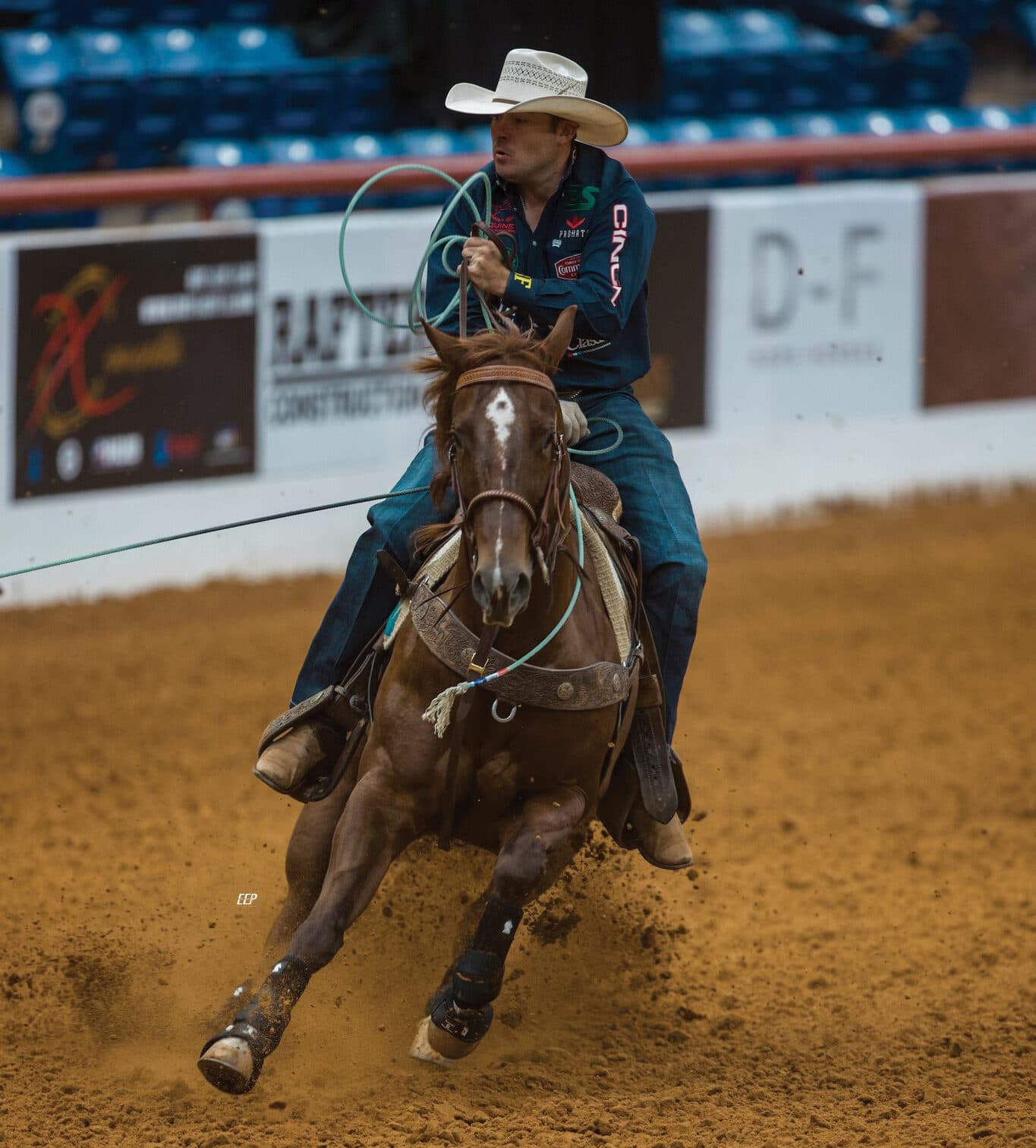 Cowboy in a white hat and teal shirt swings his rope as his sorrel horse slides hard through arena dirt