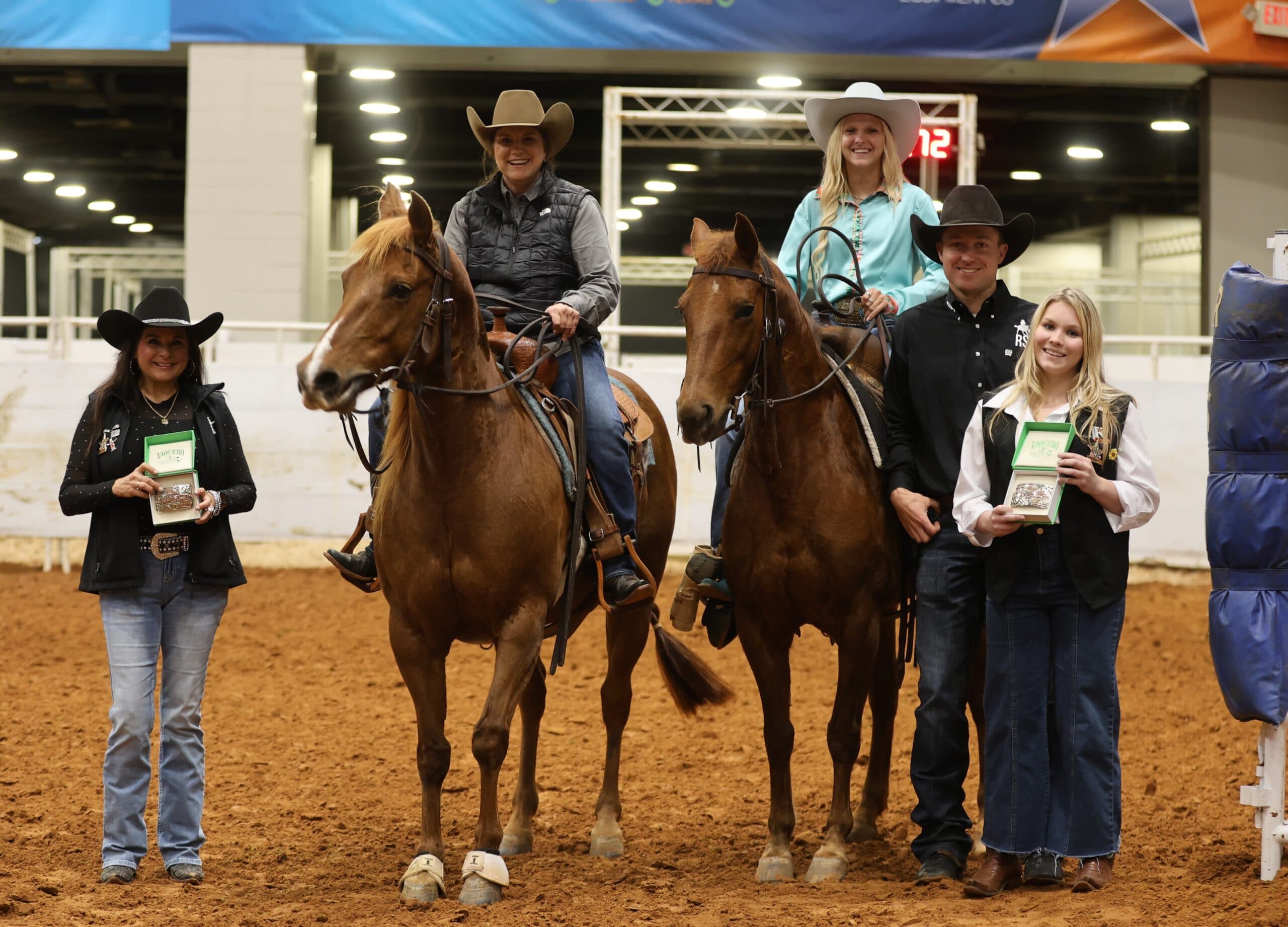 Two riders on sorrel horses pose in an arena beside three people in cowboy hats holding boxed trophy buckles