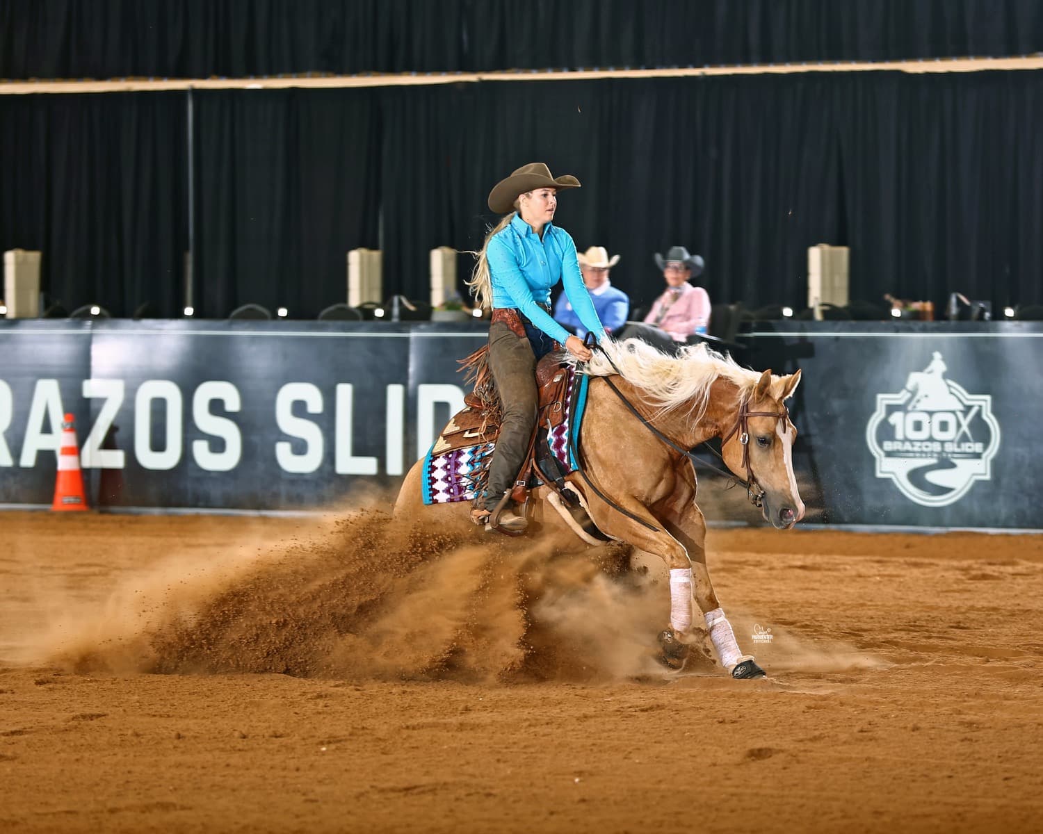 Rider in a turquoise shirt and cowboy hat slides her palomino reining horse to a stop, throwing up a spray of dirt