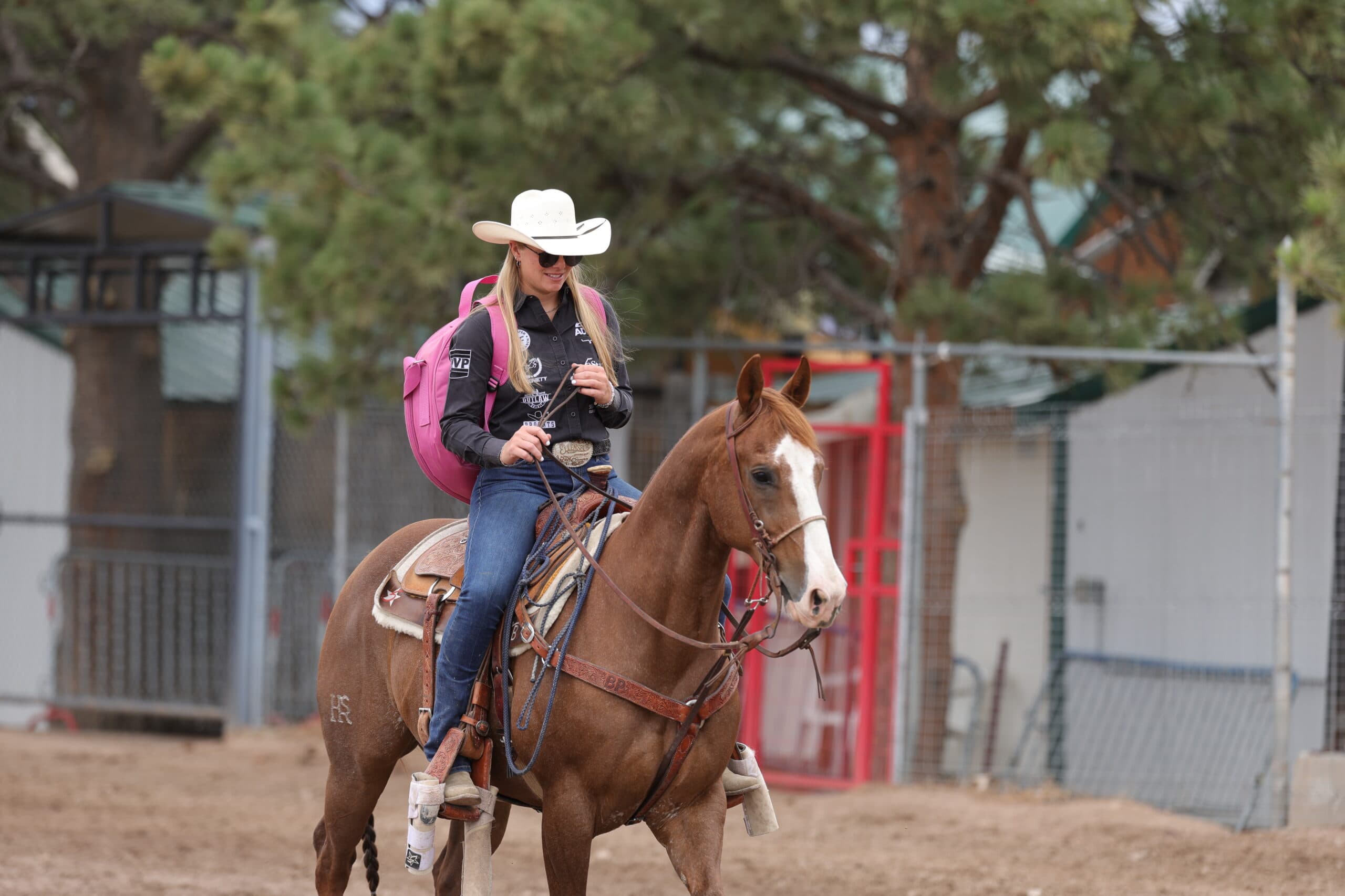 Blonde cowgirl in a white straw hat, sunglasses and pink backpack rides a sorrel horse with a blaze through a warm-up pen