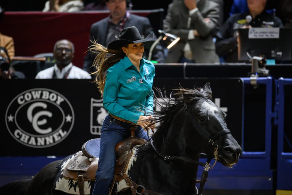 Smiling cowgirl in a teal shirt and black hat rides a dark horse past the Cowboy Channel arena banner