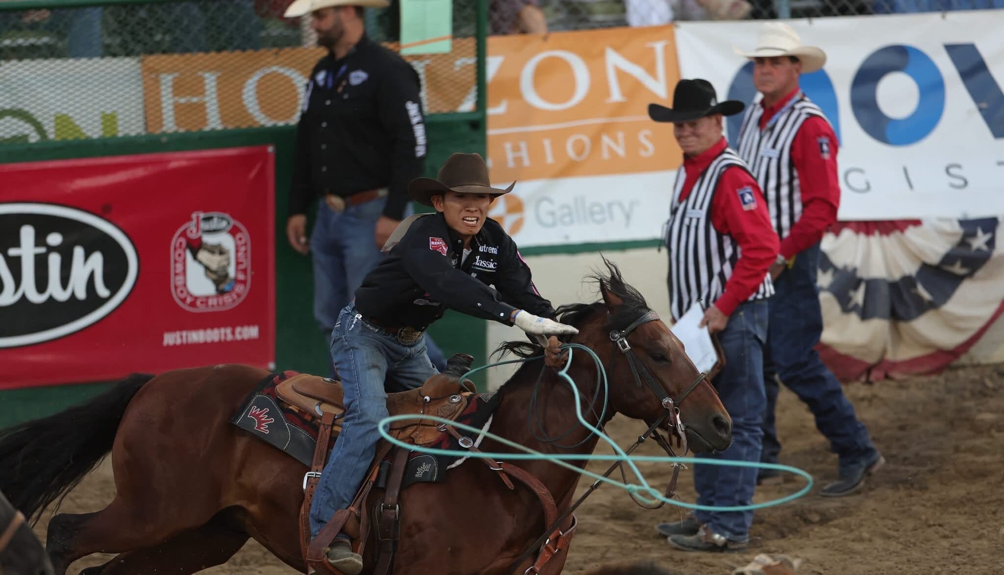 Cowboy on brown horse swinging rope during breakaway roping event at indoor rodeo arena with judges and spectators watching