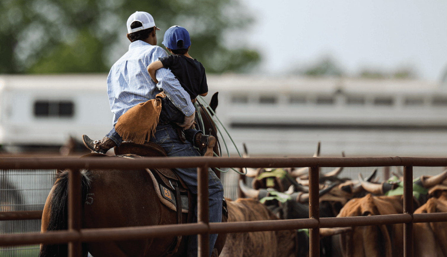 Cowboy on horseback holding a child, both wearing caps, positioned at a ranch fence with cattle visible in the background
