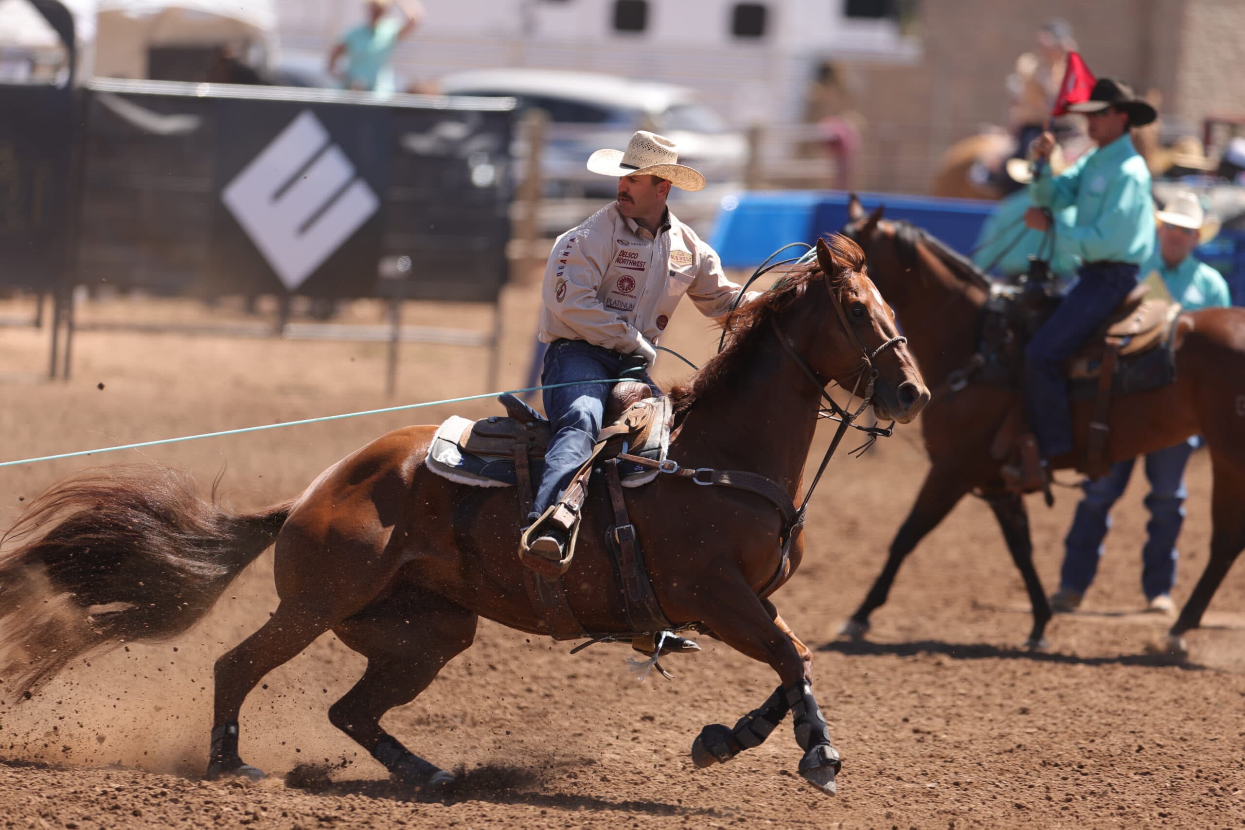 Cowboy on brown horse performing calf roping at rodeo arena, holding rope while horse moves forward in dust