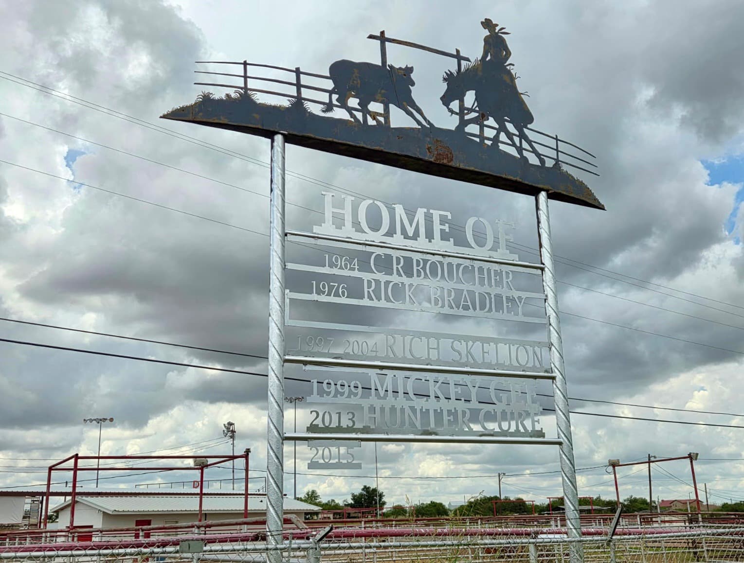 Large rodeo arena sign reading "HOME OF" with silhouetted cowboy and cattle figures on top, listing champion names and years from 1964 to 2015