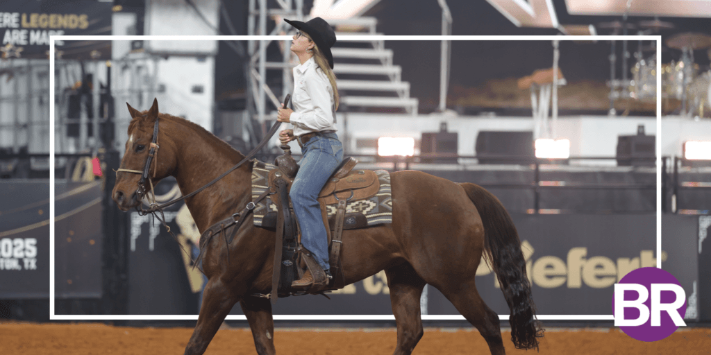 Cowgirl in a black hat and white shirt rides a sorrel horse across the arena dirt at an indoor rodeo