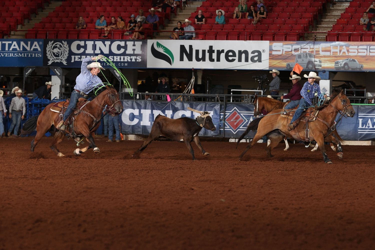 Team ropers on horseback work a steer during competition at an indoor rodeo arena with sponsor signage visible along the perimeter