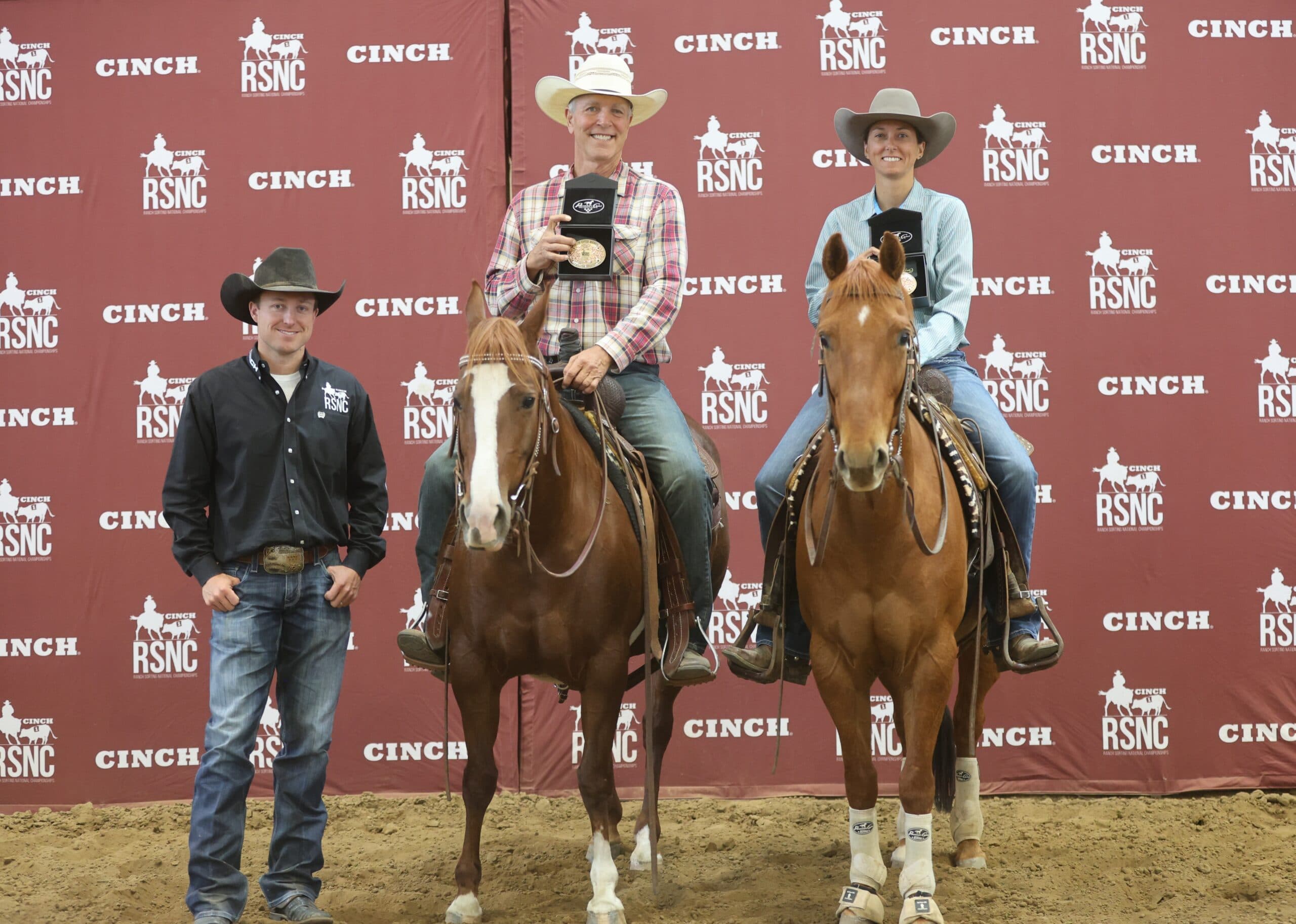 Two mounted riders hold up championship buckles beside a man standing on the ground in front of a Cinch RSNC backdrop