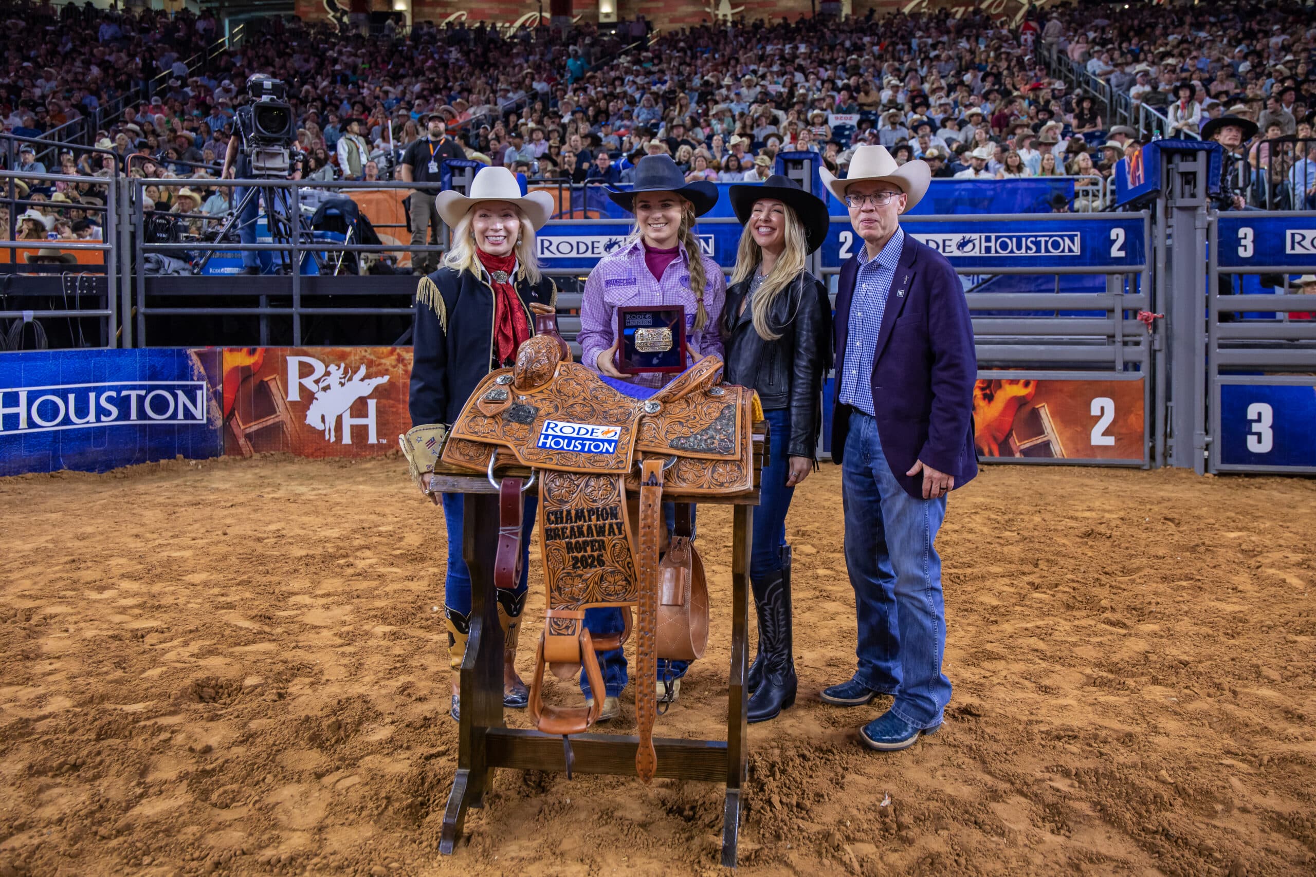 Breakaway roping champion holds a RodeoHouston buckle beside a tooled trophy saddle with three presenters in the arena
