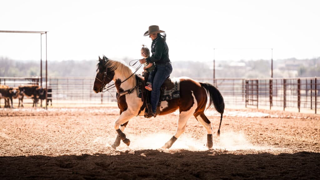 Woman in a cowboy hat rides a paint horse across a dusty arena with a small child seated in front of her