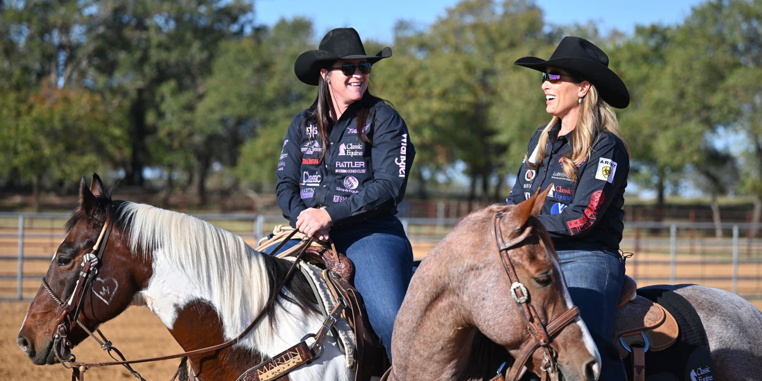 Two women in black cowboy hats and sponsor-covered shirts smile while sitting horseback in a sunny outdoor arena