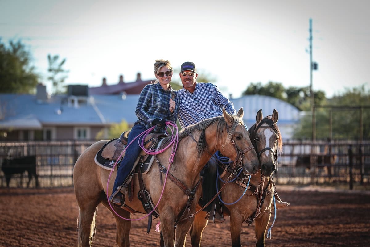 Smiling couple sit side by side horseback in a dirt arena, each carrying a coiled rope on their saddle