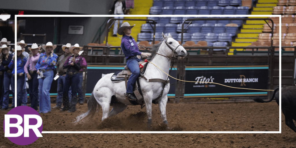 Breakaway roper in a purple shirt and cowboy hat rides a gray horse as her rope stretches toward a calf in the arena