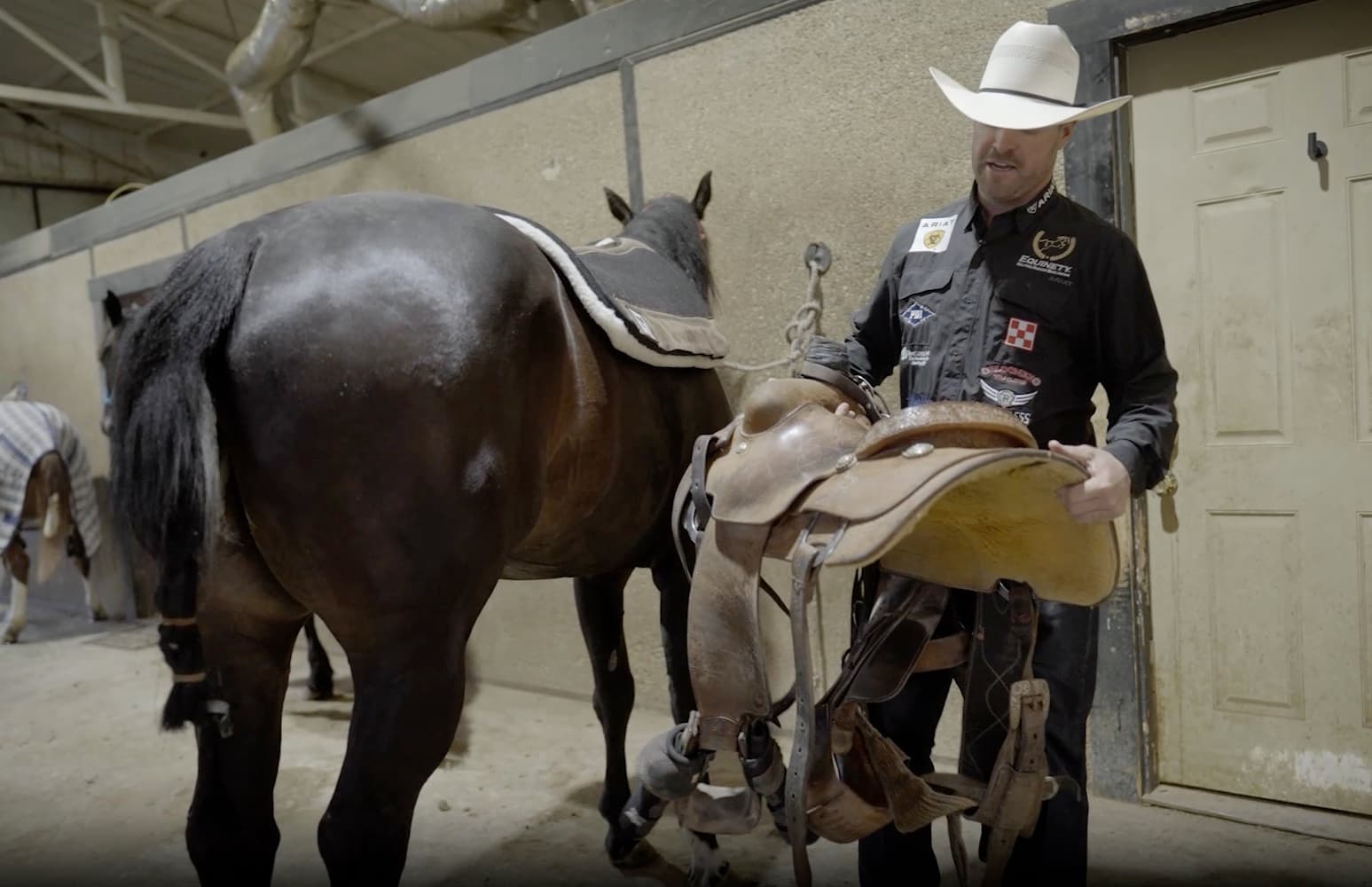 Cowboy in a white straw hat and sponsor shirt lifts a western roping saddle toward a dark horse in a barn aisle