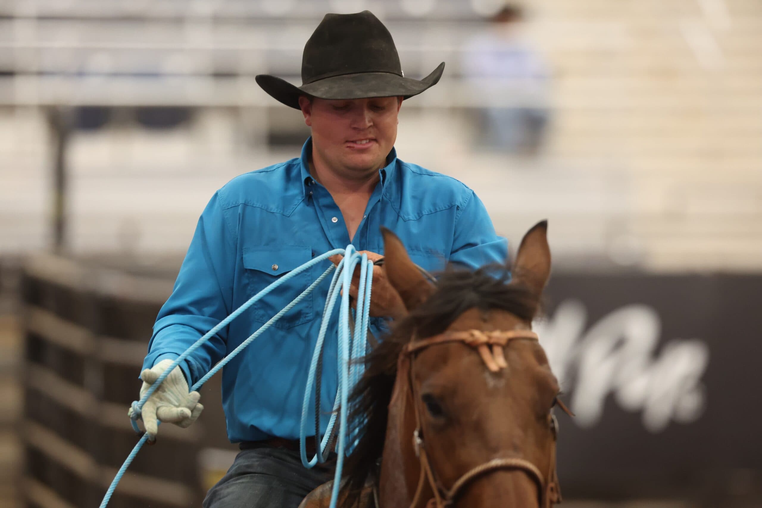 Cowboy in a black hat and blue shirt coils his light blue rope while riding a sorrel horse in the arena