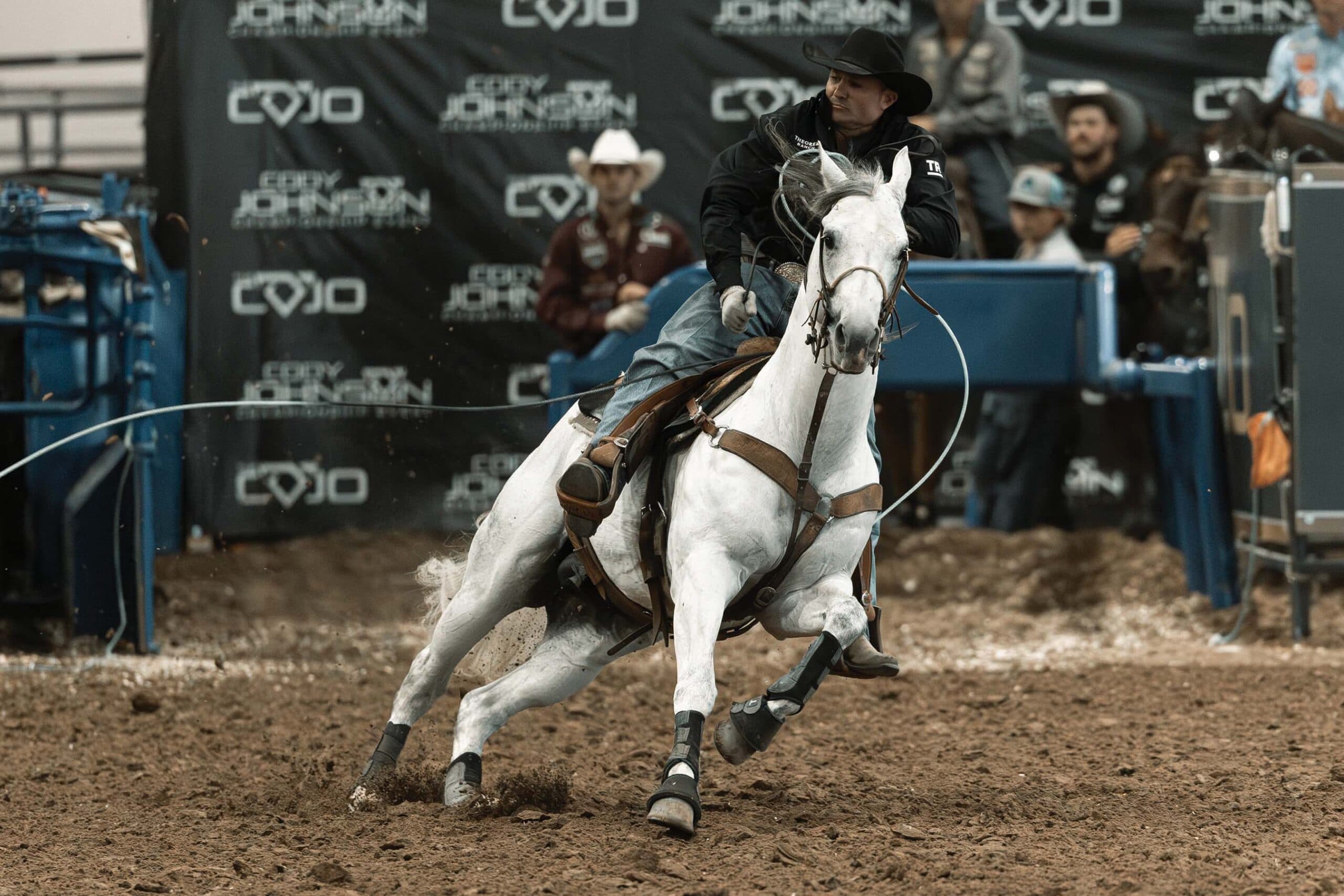 Roper in black jacket and cowboy hat rides a gray horse hard into the arena dirt, rope trailing behind
