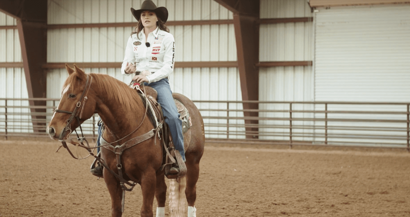 Rider in a black cowboy hat and white sponsor shirt sits astride a sorrel horse in a covered arena, speaking to the camera