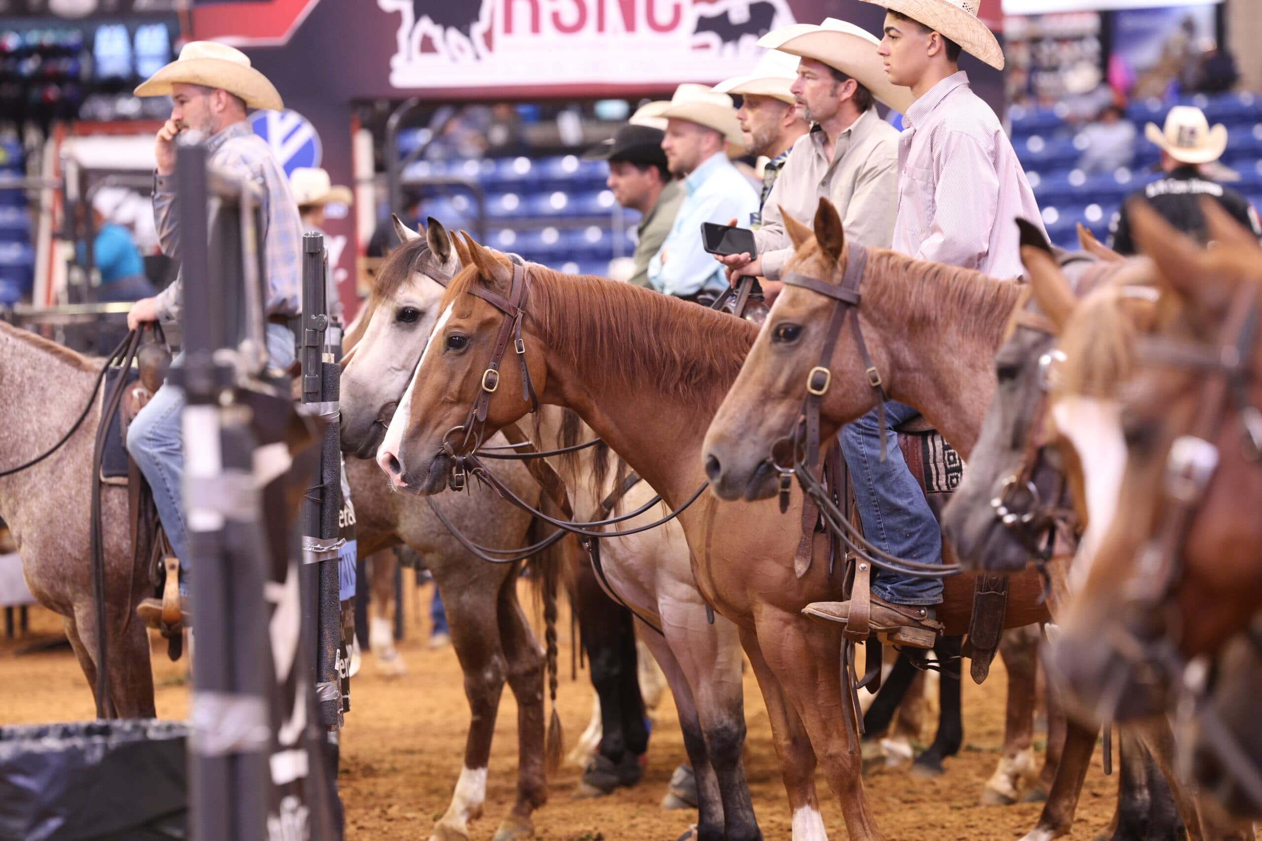 Row of cowboys in straw hats sit horseback along the arena fence beneath an RSNC banner, waiting their turn
