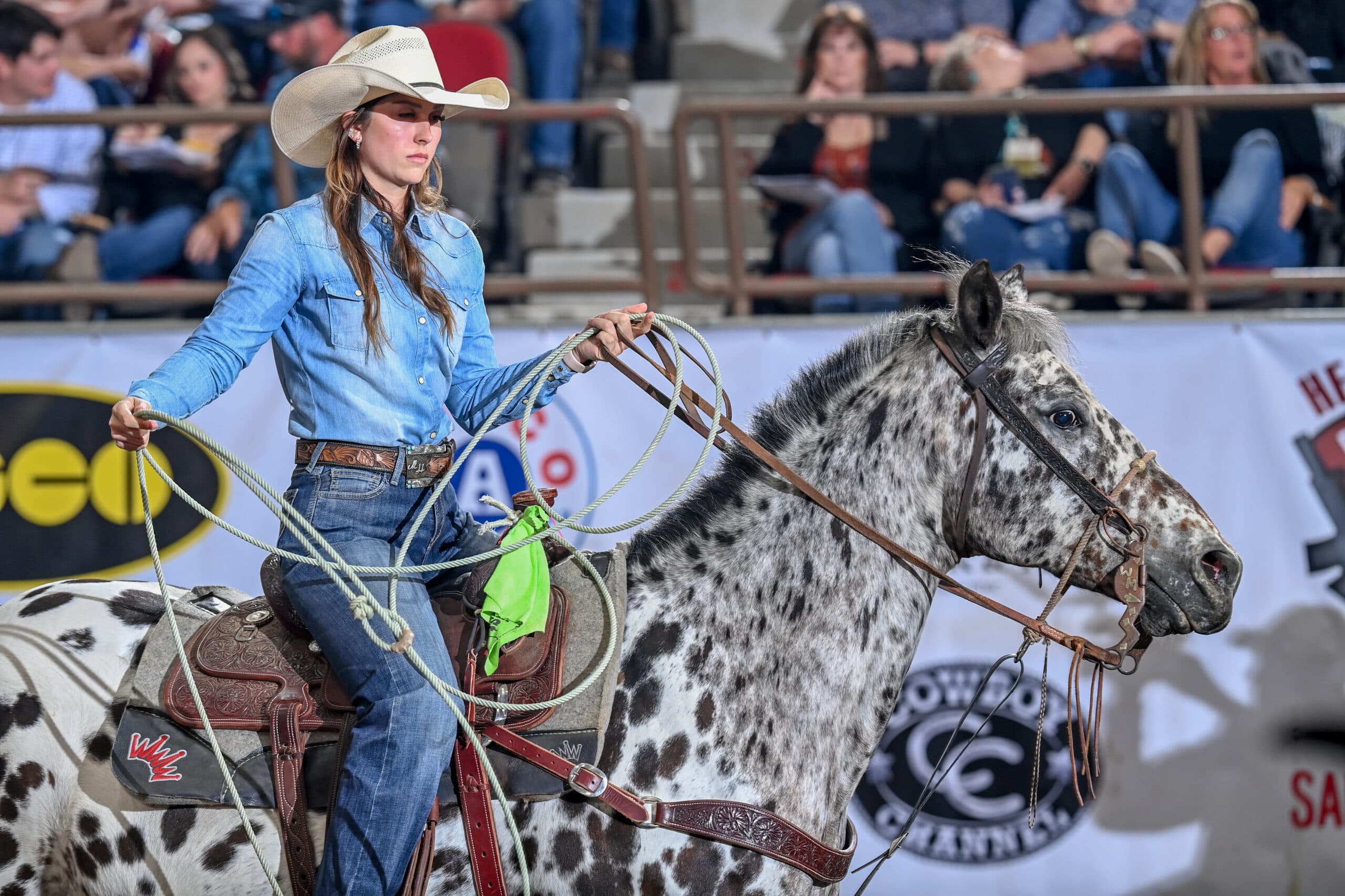 Cowgirl in a straw hat and denim shirt swings her rope while riding a spotted Appaloosa in an indoor arena