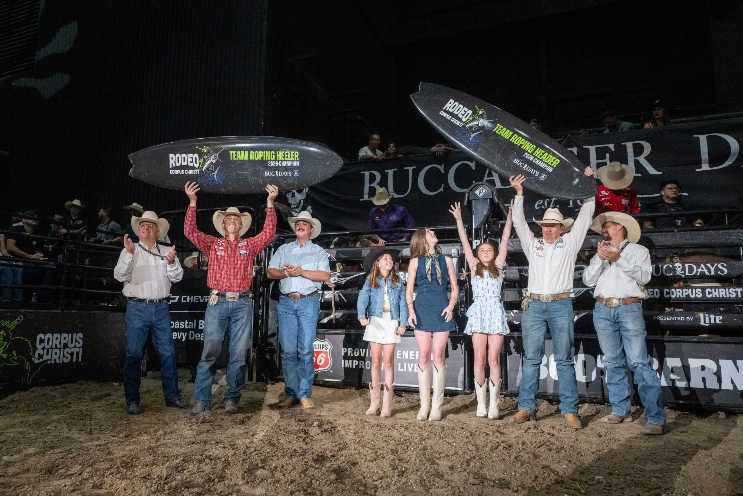 Team Roping Heeler 2020 Champion holds surfboard-style award aloft while posing with family and fellow competitors at Corpus Christi rodeo