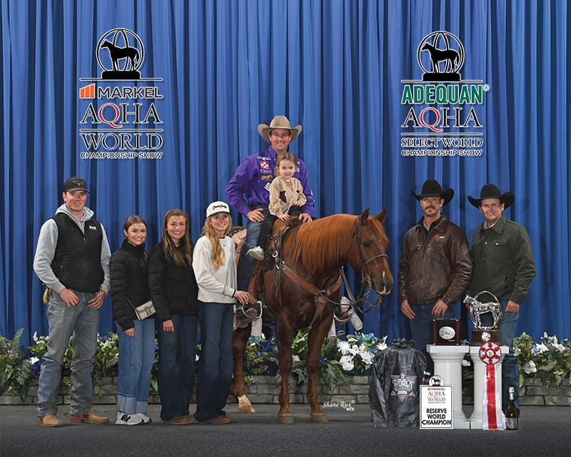 Rider in a purple shirt holds a child aboard a sorrel horse as family and friends gather for an AQHA World Show Reserve World Champion win photo