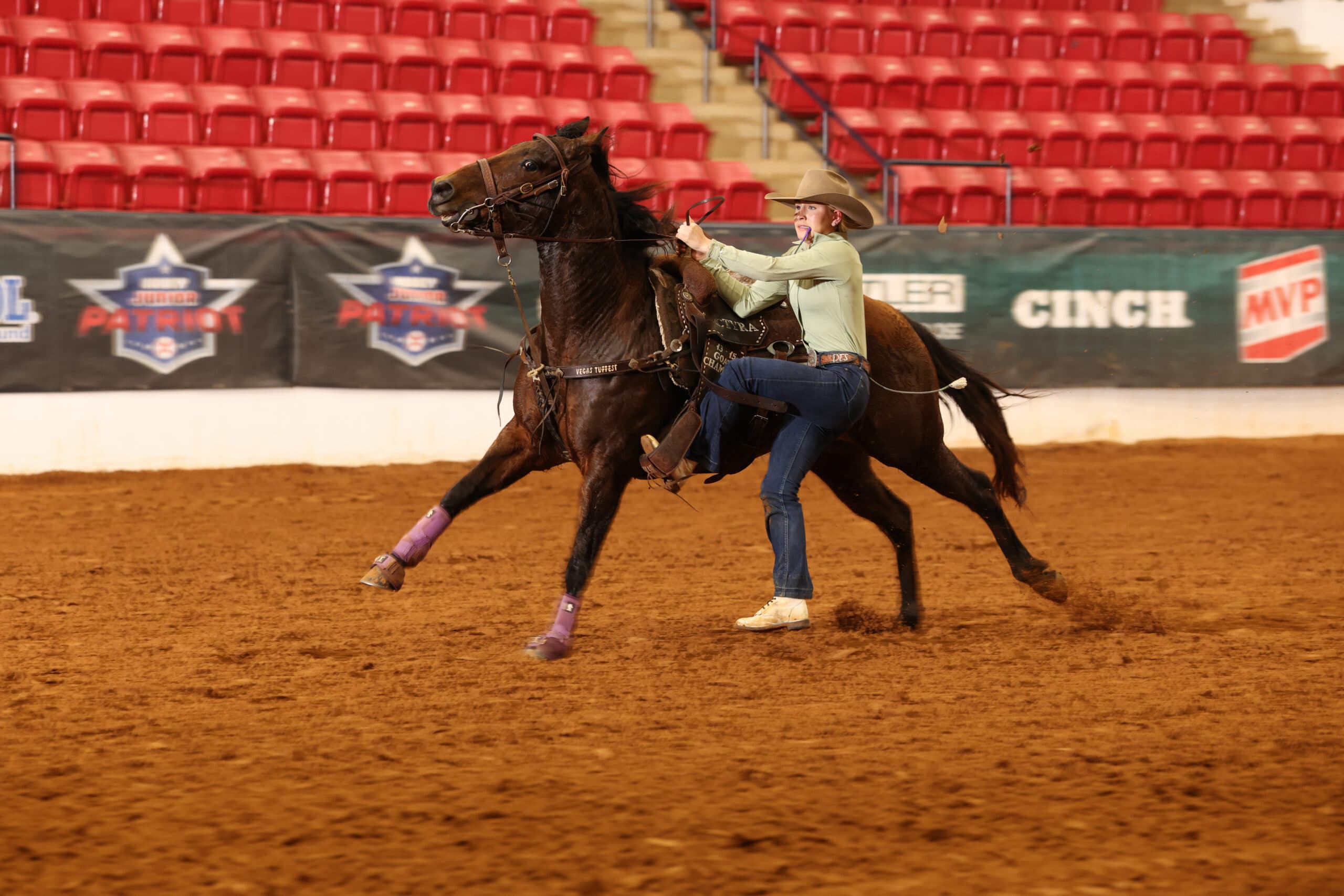 Goat tier competitor dismounting at a run from her sliding bay horse in an indoor arena with red seats behind