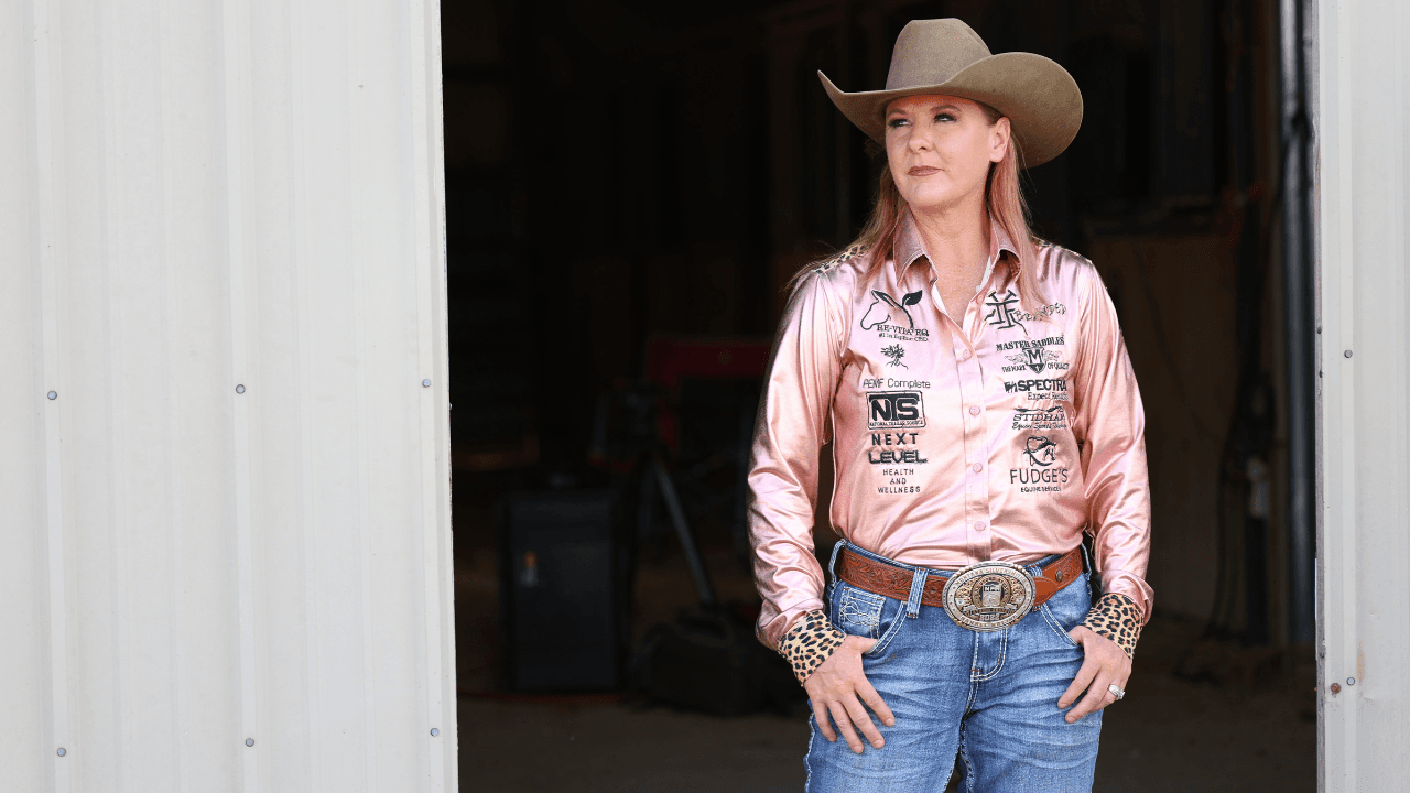 Cowgirl in a tan cowboy hat and pink sponsor shirt with a trophy buckle stands in a barn doorway