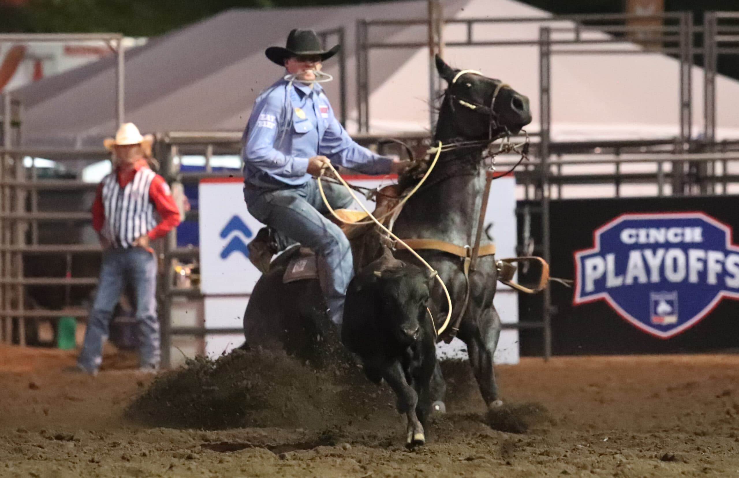 Header roper in blue shirt sits deep as his dark horse stops hard, rope tight on a black steer in the arena dirt