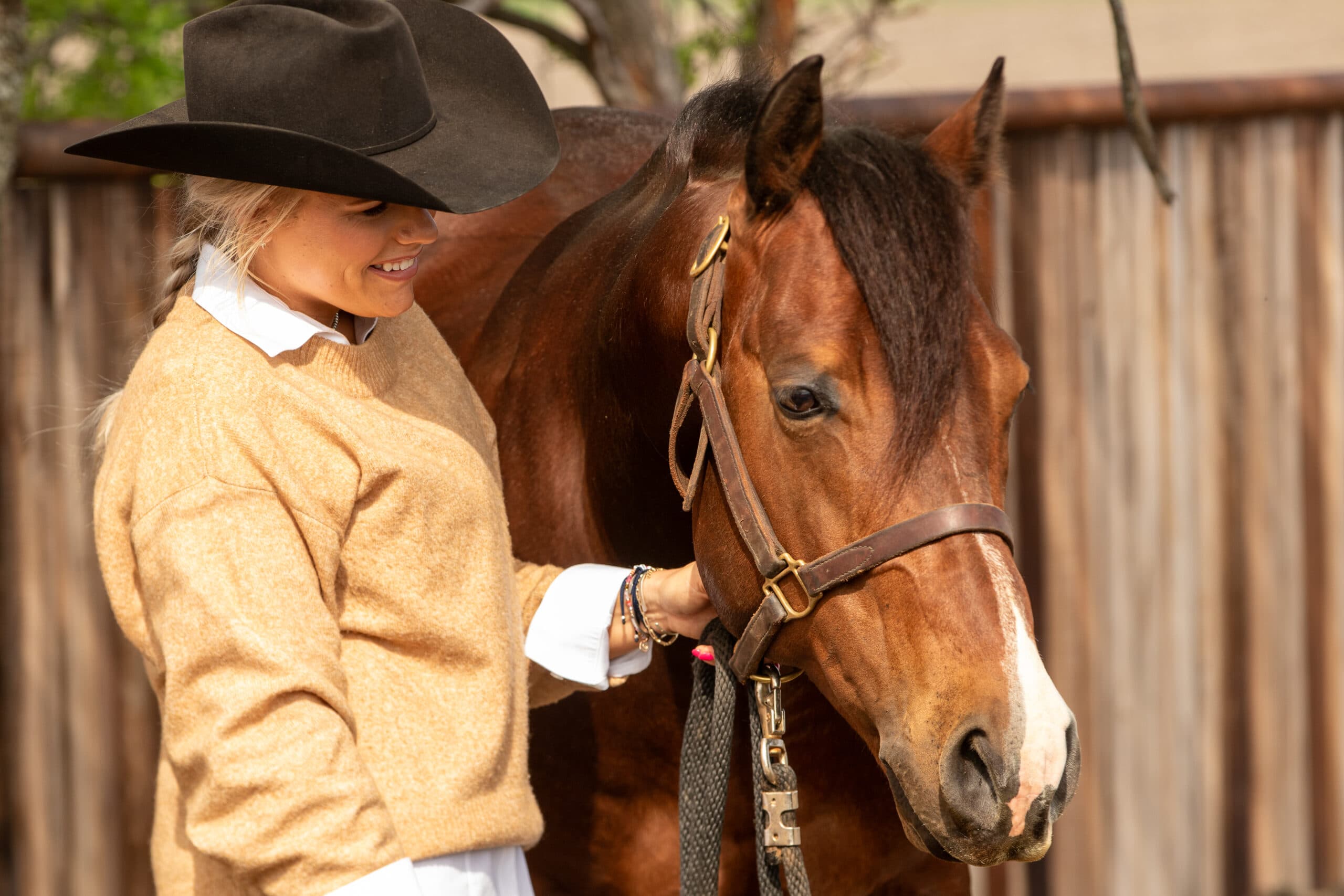 Cowgirl in tan shirt and black cowboy hat smiles while holding the halter of a bay horse with white blaze at a ranch fence