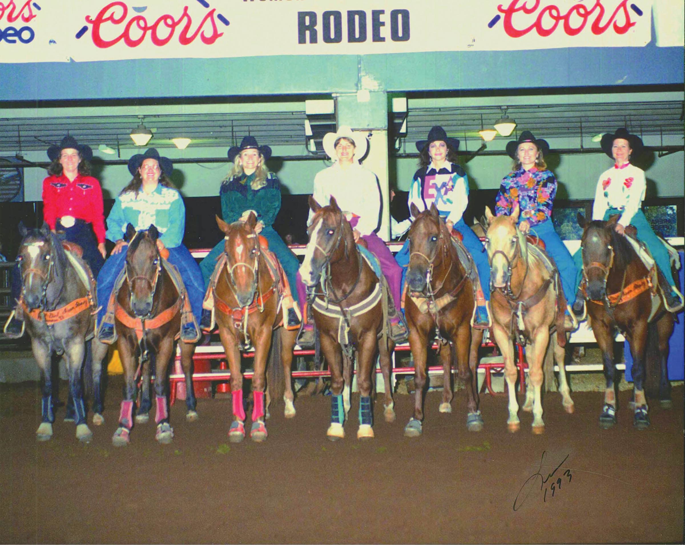 Seven cowgirls on horseback line up side by side in an arena beneath Coors rodeo banners, 1993
