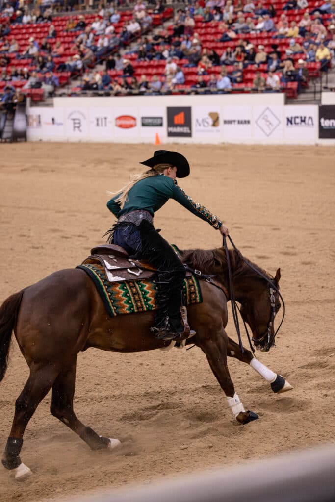 Rider in a black cowboy hat and teal shirt slides her sorrel horse to a stop in the arena as spectators watch from the stands
