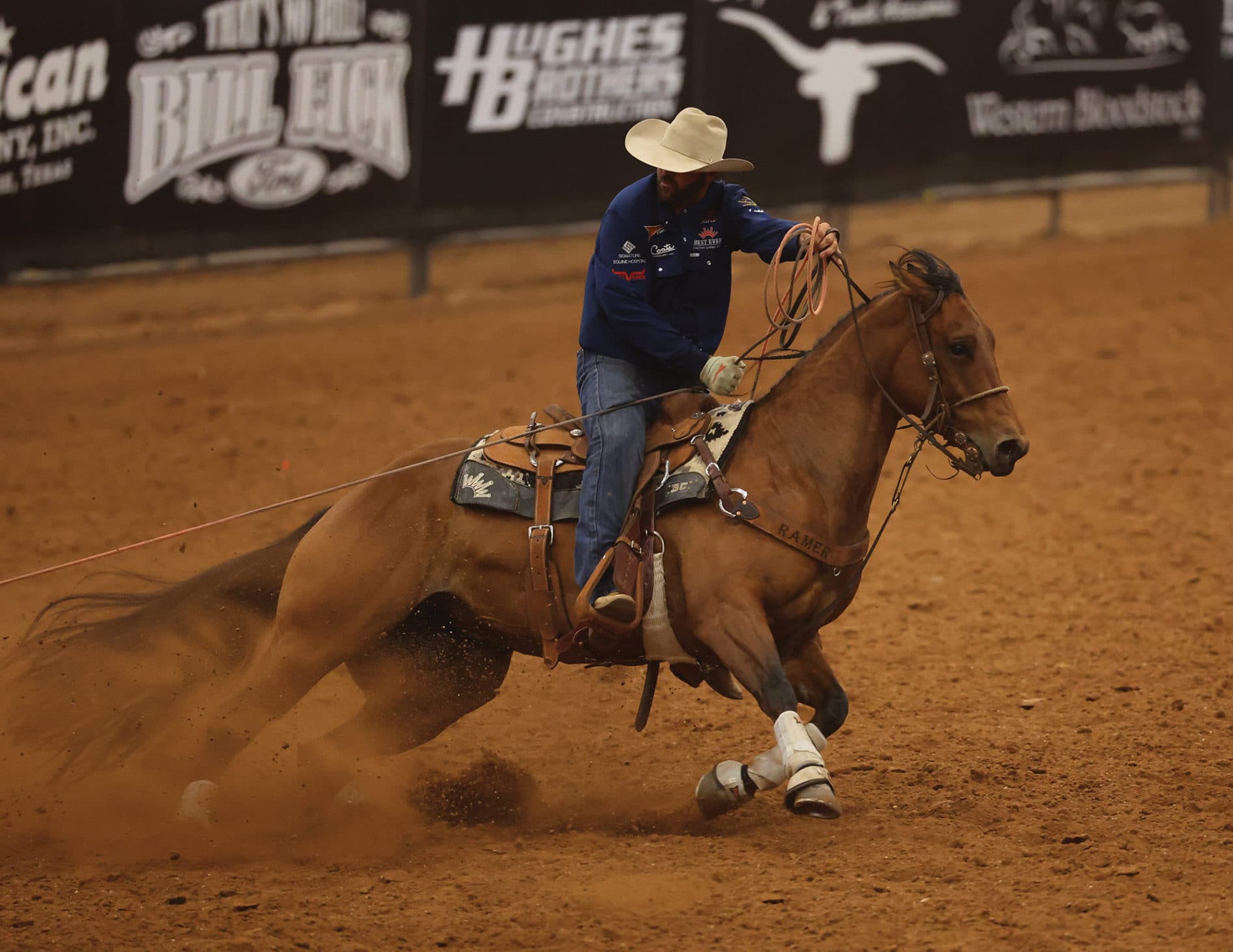 Header in blue shirt and straw cowboy hat rides a buckskin horse hard into the dirt as the taut rope stretches behind them