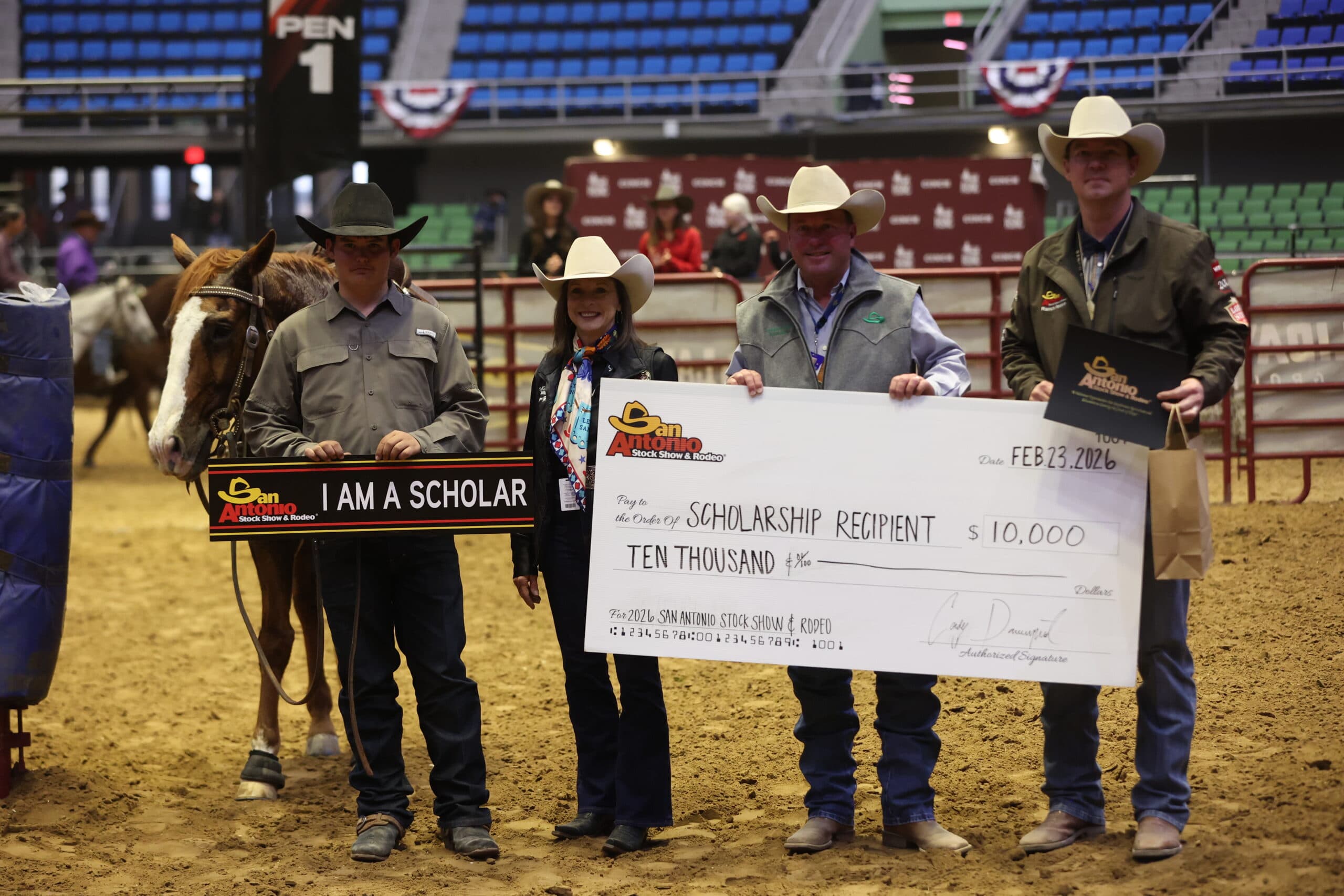 Scholarship recipient holding a horse and "I Am A Scholar" sign beside three people with a giant $10,000 San Antonio Stock Show & Rodeo check in the arena