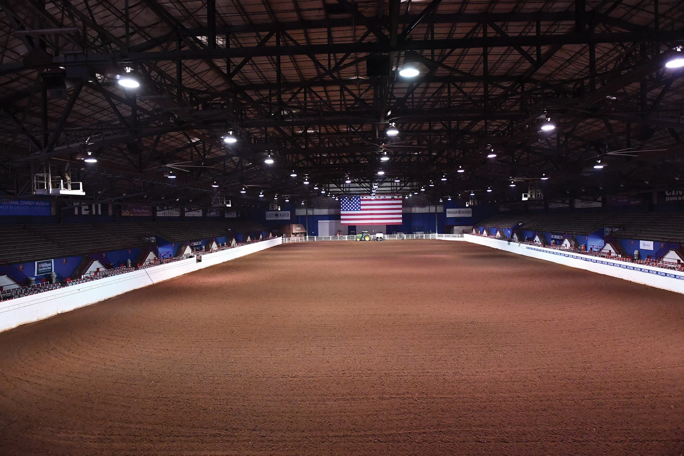 Empty indoor rodeo arena with dirt floor, white rail fencing, American flag backdrop, and stadium seating under industrial lighting