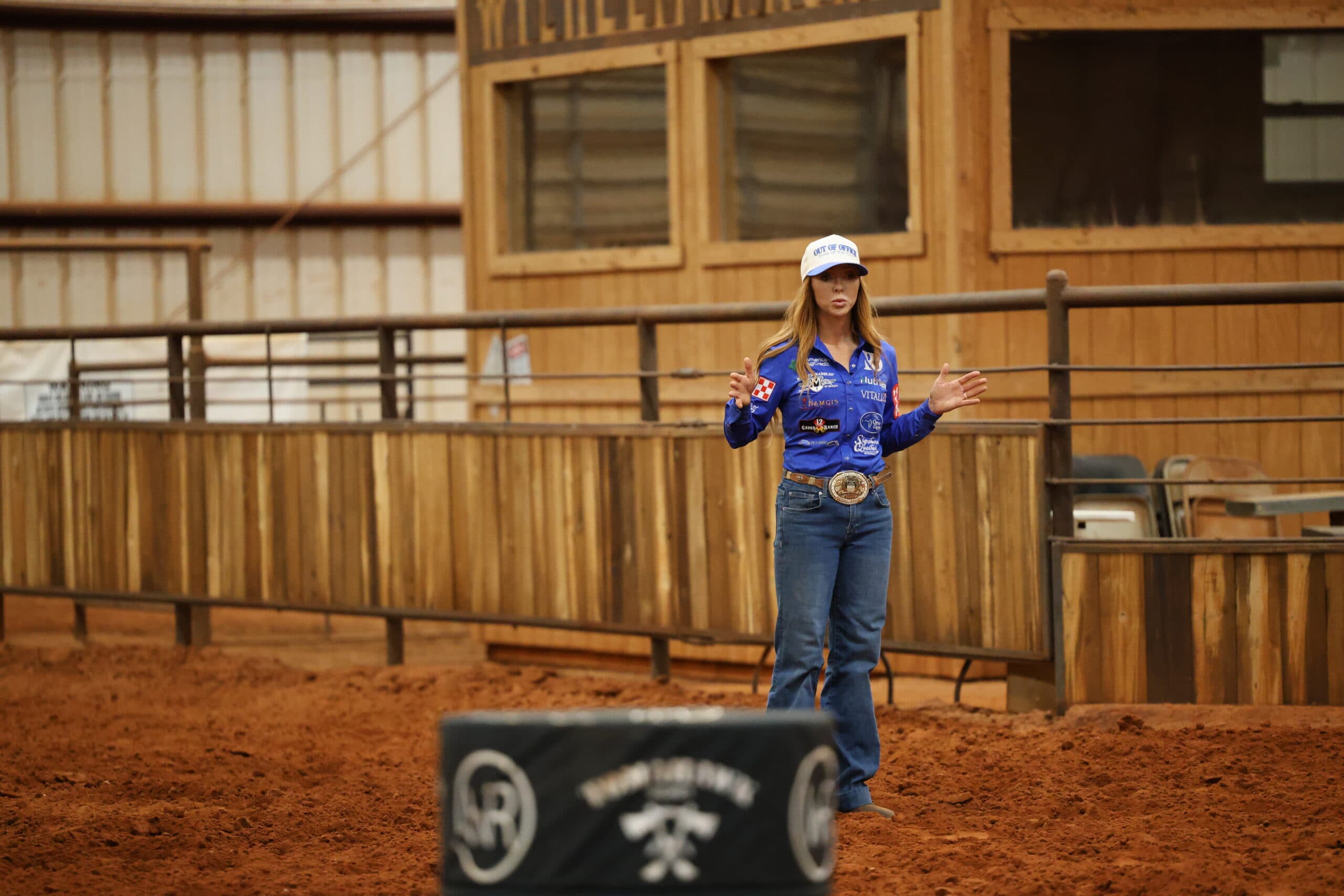Woman in a blue sponsor shirt and ball cap gestures with both hands while speaking in a dirt arena beside a padded barrel