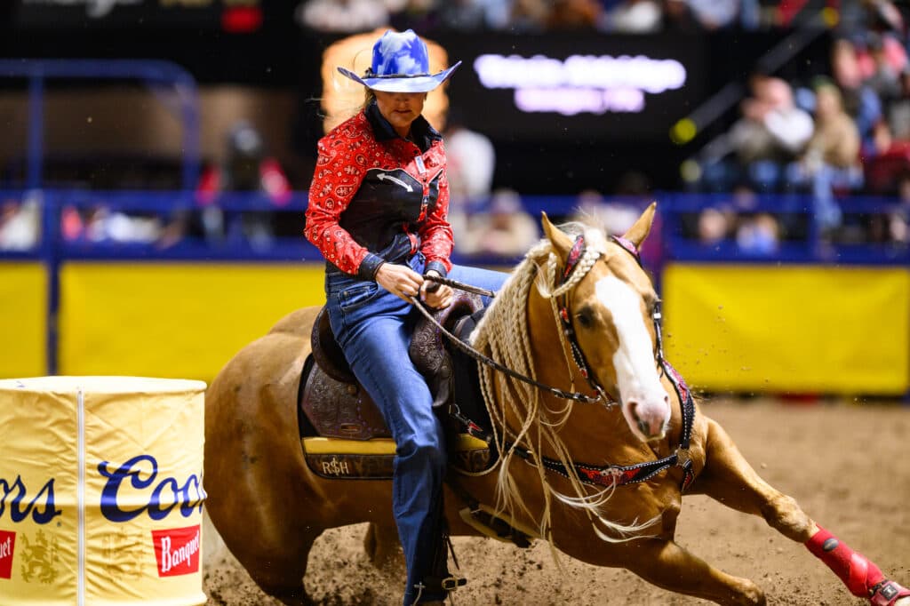 Barrel racer in a blue hat and red bandana shirt turns her palomino horse tight around a Coors Banquet barrel