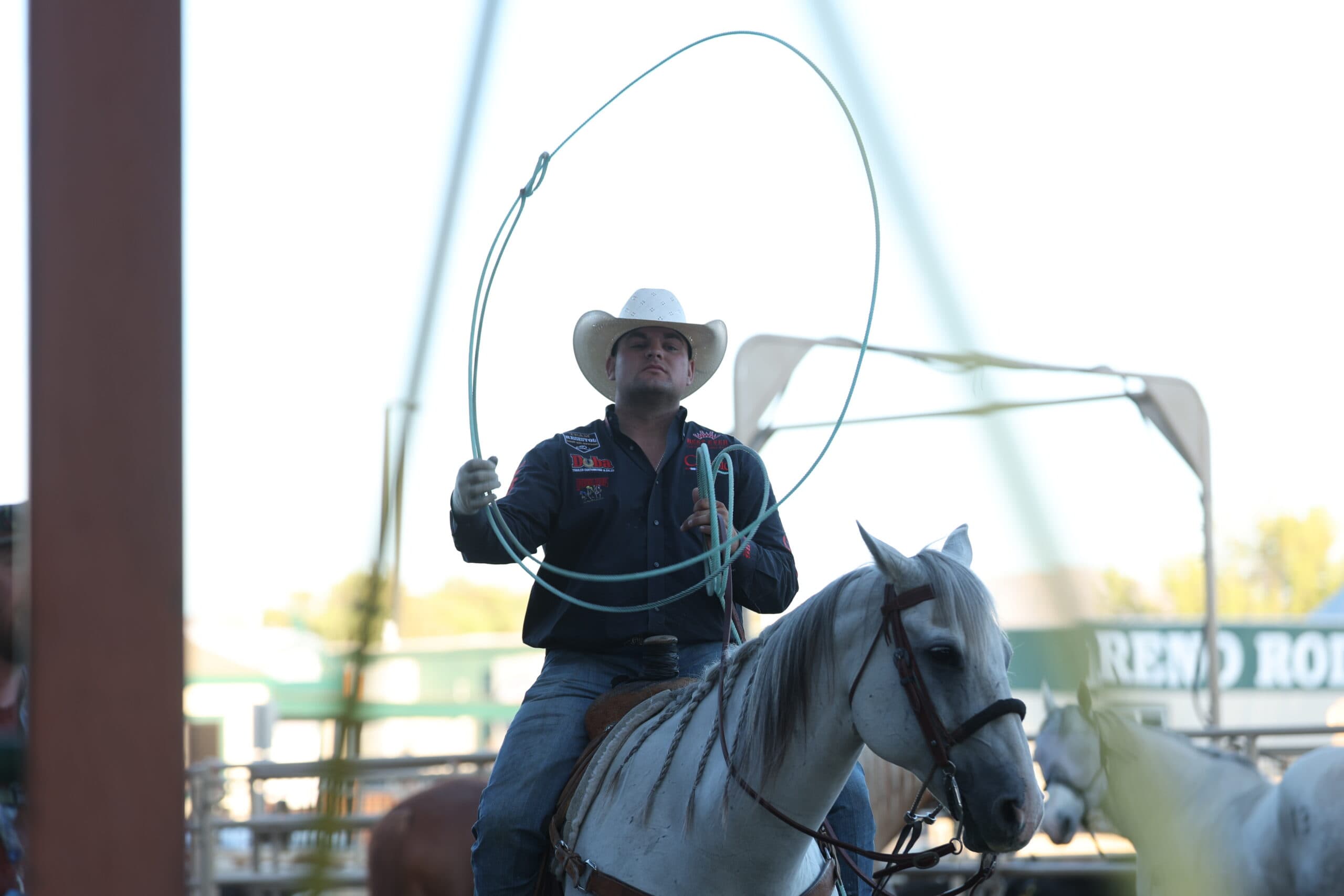 Roper on white horse swinging lasso overhead during rodeo competition in arena