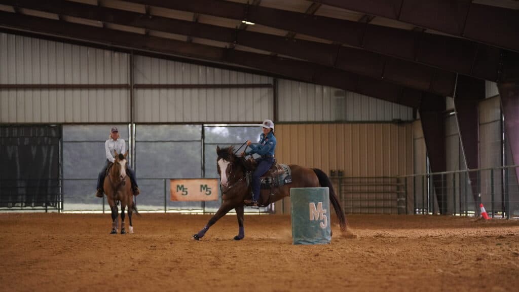 Rider in a helmet turns her bay horse around a teal M5 barrel in a covered arena as a mounted instructor watches