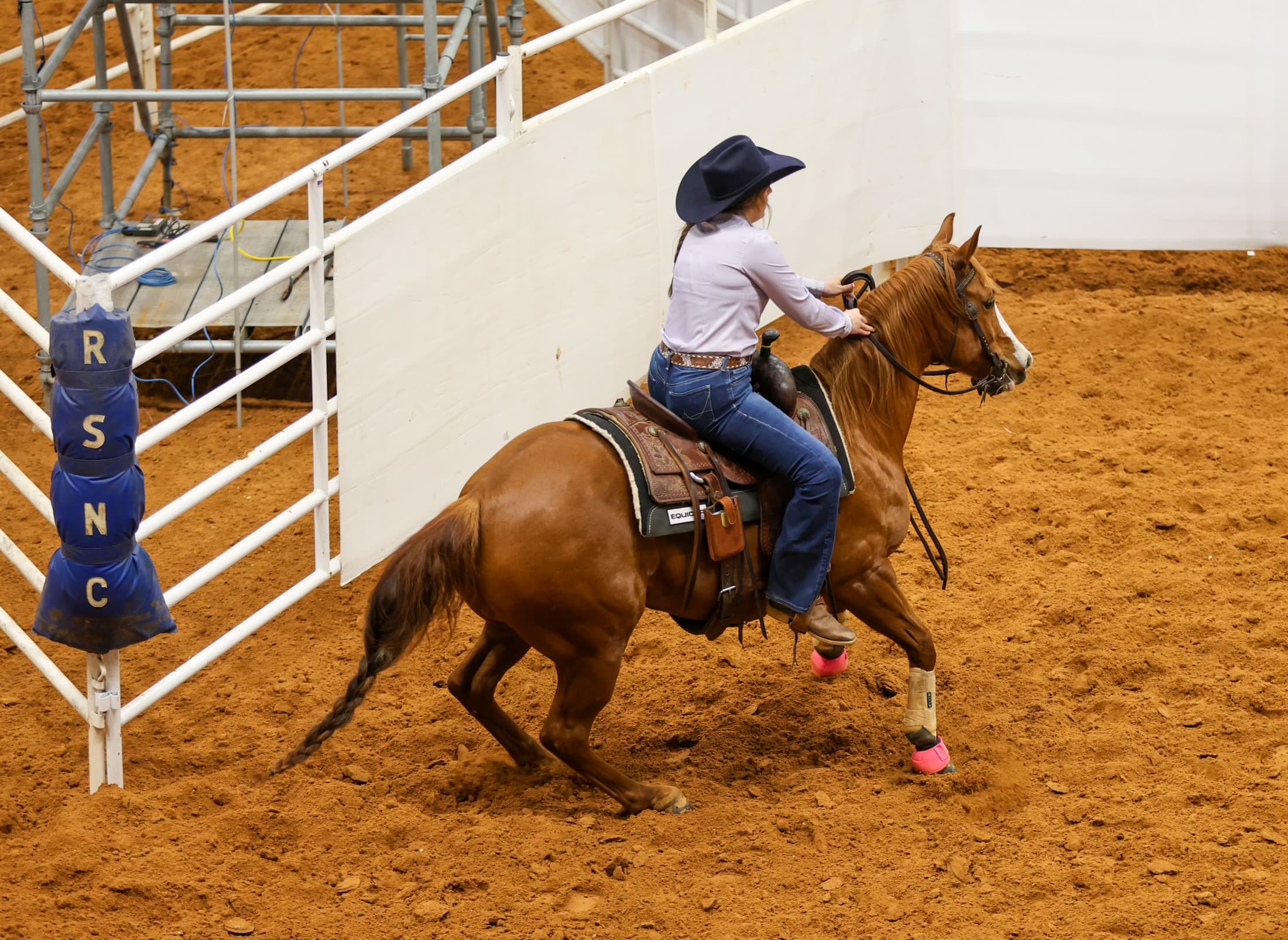 Cowgirl in a black hat rides a sorrel horse in pink hoof boots along the arena wall past an RSNC-branded gate post