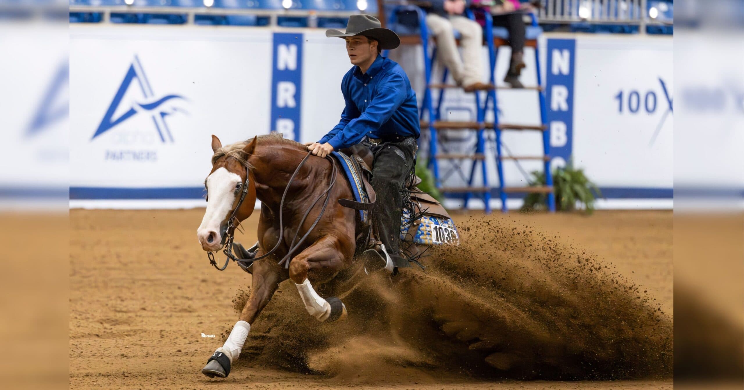 Reining horse in a sliding stop throwing up a spray of dirt as its rider in a blue shirt and black hat looks ahead