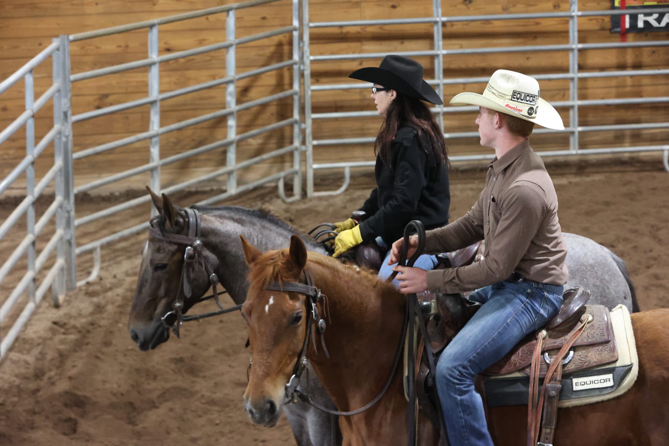 Two riders in cowboy hats sit horseback beside metal panels in an indoor arena, watching the action outside the frame